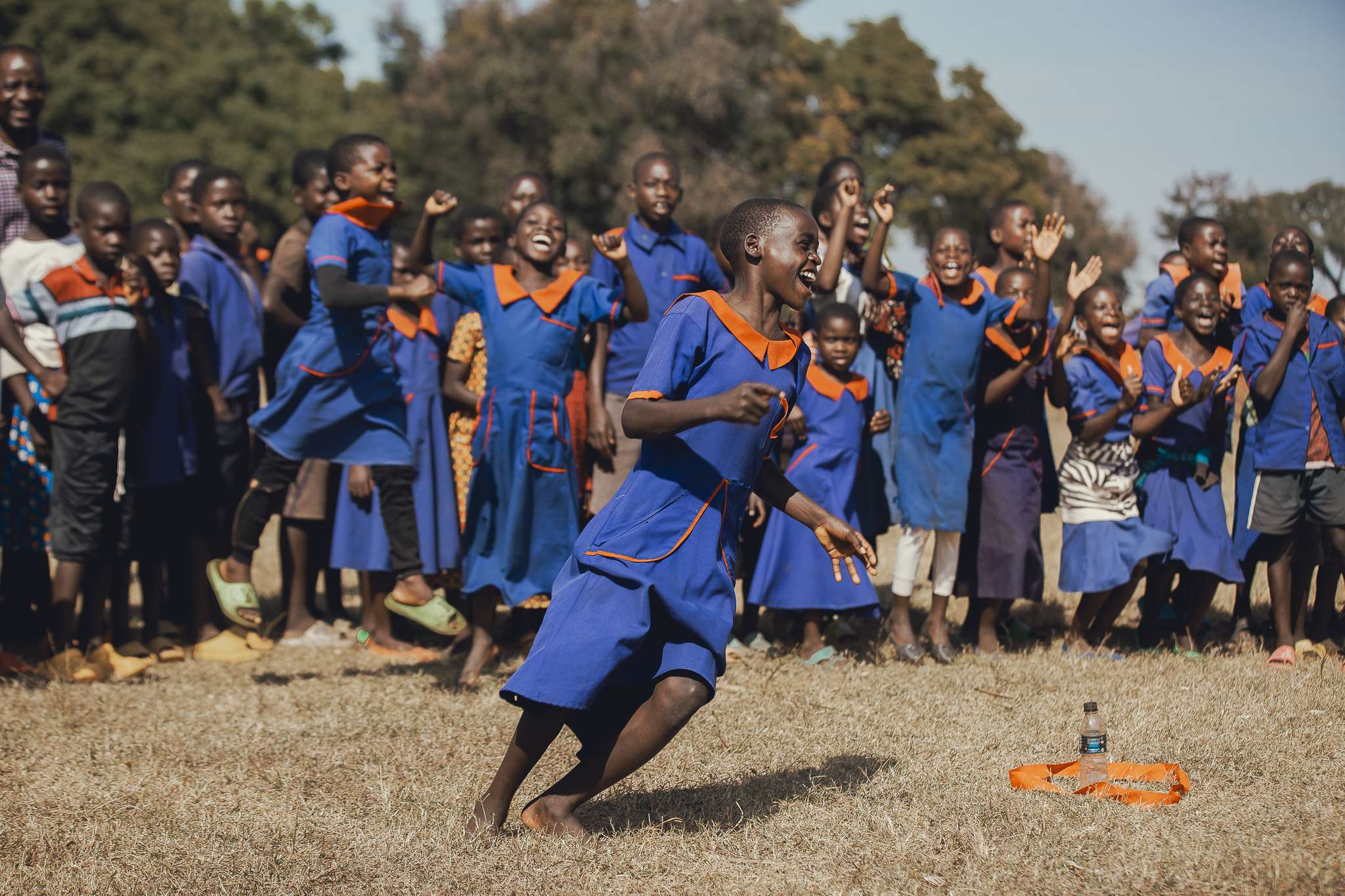 A teacher and a group of smiling schoolchildren playing together outdoors in East Africa.