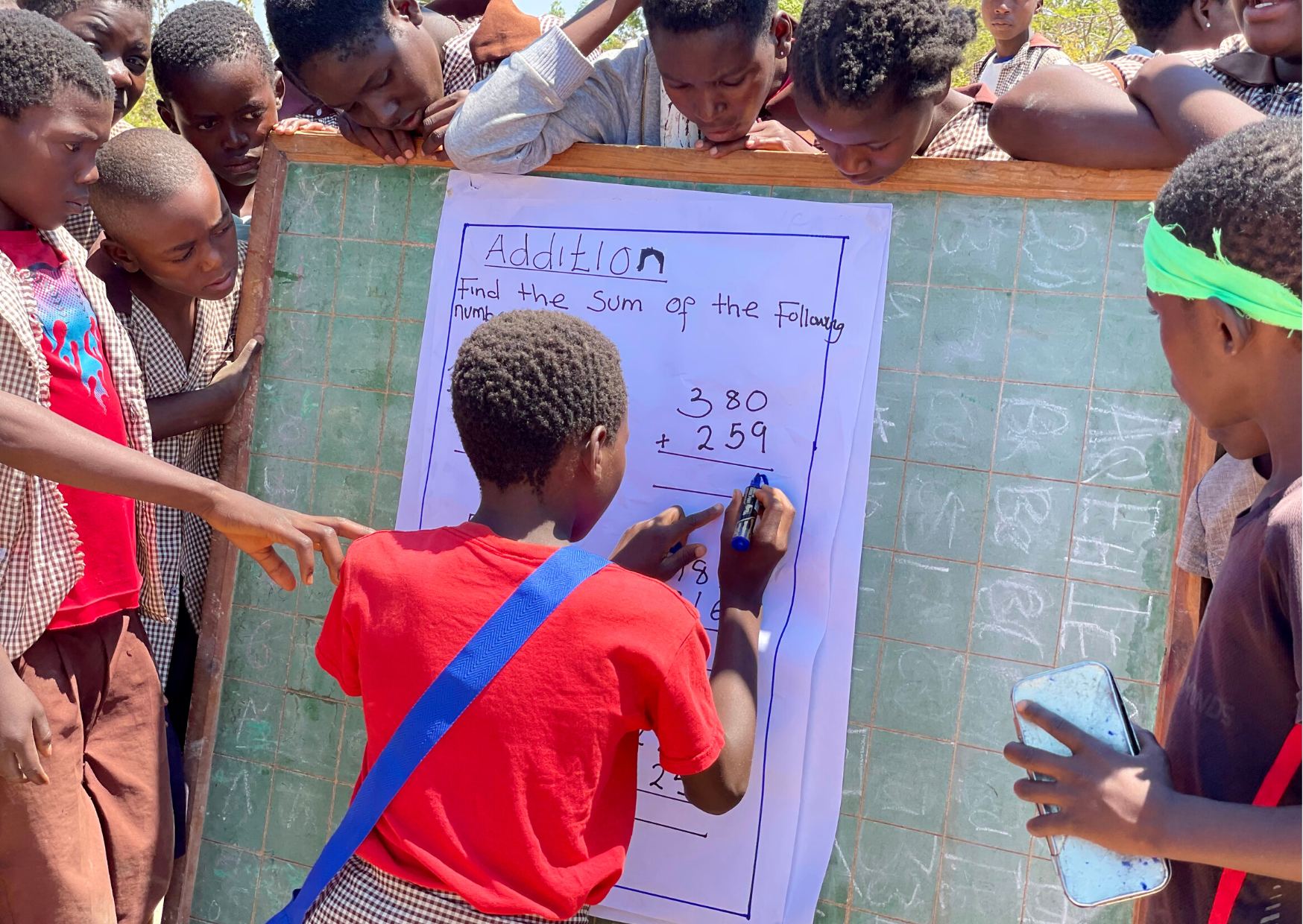 Children crowding around a flipchart on a blackboard, working out an addition sum.