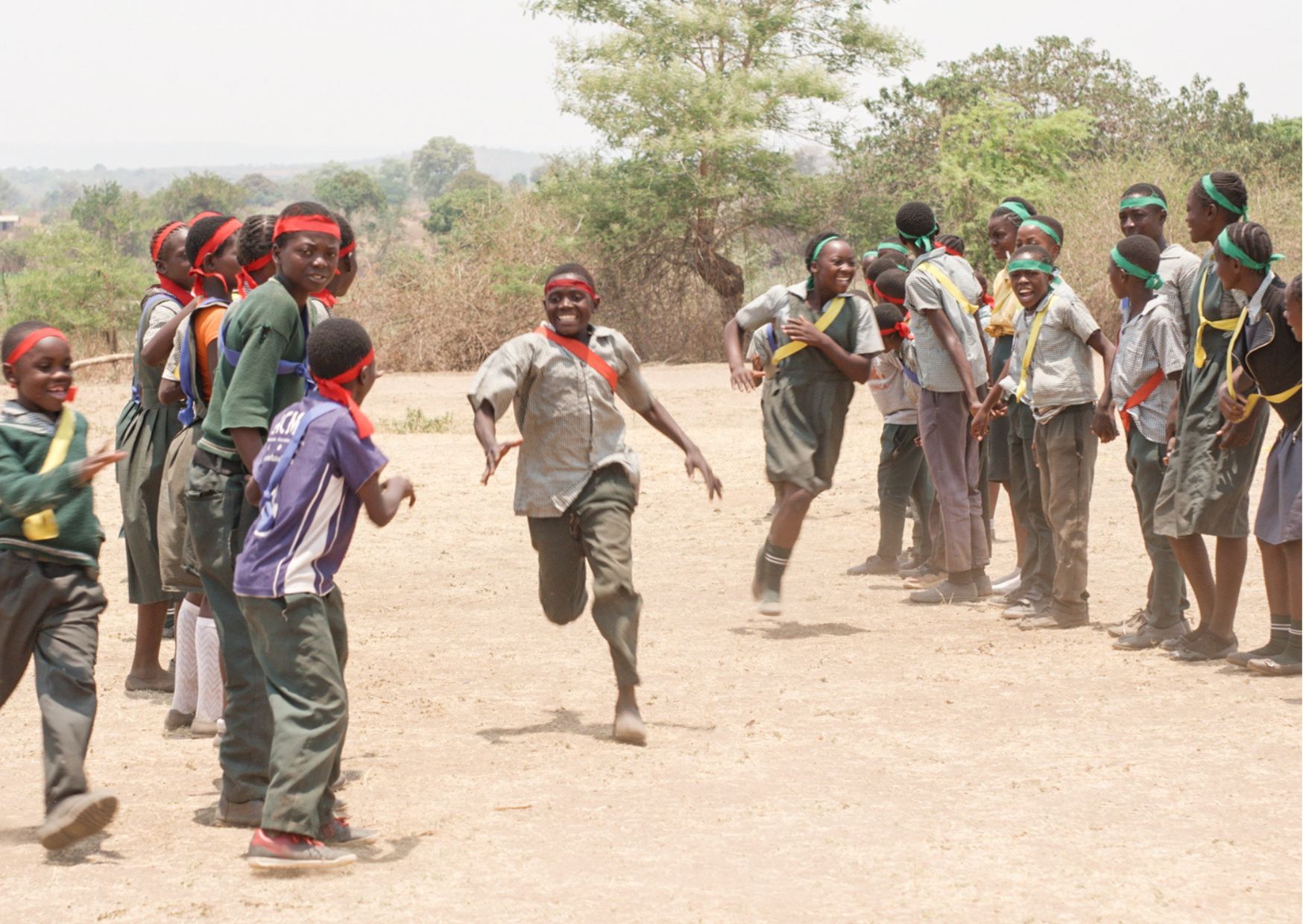 A child running across a dusty field while classmates wait along the side.